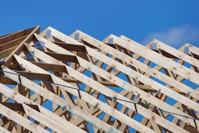 Wooden roof trusses arranged in a triangular pattern against a clear blue sky. Metal connectors hold the beams together, indicating the early stage of construction.