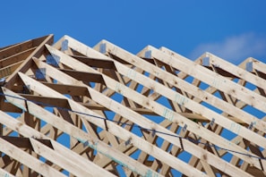 Wooden roof trusses arranged in a triangular pattern against a clear blue sky. Metal connectors hold the beams together, indicating the early stage of construction.