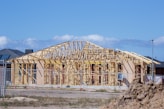 An unfinished house structure with wooden framing is surrounded by a construction site. The frame outlines the shape of the house, indicating rooms and the roof. There's a dirt mound in the foreground, a fence surrounding the construction area, and other houses in the background under a clear blue sky.