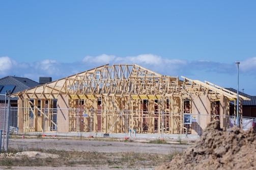 An unfinished house structure with wooden framing is surrounded by a construction site. The frame outlines the shape of the house, indicating rooms and the roof. There's a dirt mound in the foreground, a fence surrounding the construction area, and other houses in the background under a clear blue sky.