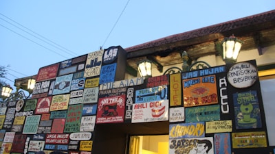 A wall covered with numerous colorful signboards, each with various writings and designs. The signs contain advertisements and information for different services such as shoe repair, rain jackets, and food vendors. The wall is illuminated by traditional street lamps, casting a warm glow against the early evening sky.