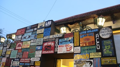A wall covered with numerous colorful signboards, each with various writings and designs. The signs contain advertisements and information for different services such as shoe repair, rain jackets, and food vendors. The wall is illuminated by traditional street lamps, casting a warm glow against the early evening sky.