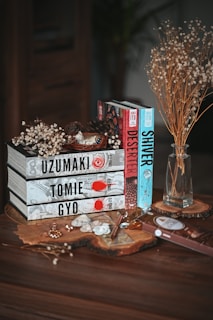 A collection of books is neatly stacked on a wooden surface. The titles are visible and include 'Uzumaki', 'Tomie', and 'Gyo', all with decorative covers. Next to them, there are more books standing upright, titled 'Deserter' and 'Shiver'. Dried flowers in a glass vase and smaller botanical arrangements rest nearby, adding a rustic touch. Additionally, there's a chocolate bar, a spoon, and some small decorative items arranged artistically.