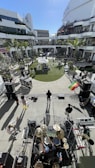 A band is performing outdoors in a modern, open-air shopping center. The view is from above and behind the drummer, capturing several musicians with various instruments like keyboards and guitars. The area is surrounded by shops and lined with palm trees, with a small lawn and a few onlookers scattered around.