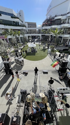 A band is performing outdoors in a modern, open-air shopping center. The view is from above and behind the drummer, capturing several musicians with various instruments like keyboards and guitars. The area is surrounded by shops and lined with palm trees, with a small lawn and a few onlookers scattered around.
