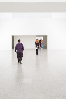 A spacious, minimalistic interior features a large, empty floor with several people walking through a doorway that leads to another section. The space has white walls and a sleek design.