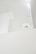A minimalistic interior view features a section of a white ceiling with a vent and lighting fixtures. The ceiling is composed of several panels, and there is a partial view of a vertical structural element.