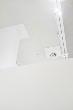 A minimalistic interior view features a section of a white ceiling with a vent and lighting fixtures. The ceiling is composed of several panels, and there is a partial view of a vertical structural element.