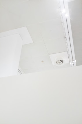 A minimalistic interior view features a section of a white ceiling with a vent and lighting fixtures. The ceiling is composed of several panels, and there is a partial view of a vertical structural element.