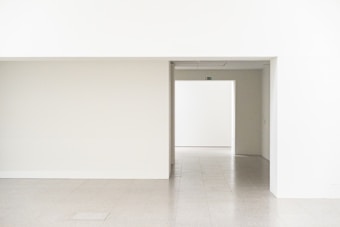 A minimalist interior setting with smooth, light-colored walls and a clean, open doorway leading into another room. The floor appears to be tiled, and the walls and ceiling are painted in a subdued white tone. An exit sign is situated above the opening, providing an element of direction.