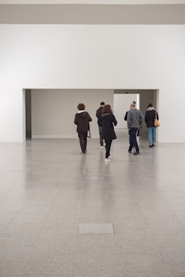A group of people walk through a minimalist, spacious gallery or museum space with a large, white wall and a small doorway in the distance. The floor is made of smooth, gray tiles, and the atmosphere is quiet and contemplative.