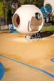 A modern playground structure features a futuristic, spherical design in a neutral, white color. The structure includes circular openings and is connected to a cylindrical tunnel made of blue netting material. The playground area has a soft, rubberized surface with decorative blue lines.