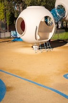 A modern playground structure features a futuristic, spherical design in a neutral, white color. The structure includes circular openings and is connected to a cylindrical tunnel made of blue netting material. The playground area has a soft, rubberized surface with decorative blue lines.