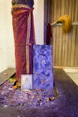 A tree trunk is wrapped in a maroon and gold patterned cloth. In front of the tree trunk, there is a blue artwork depicting a figure with multiple limbs, reminiscent of traditional or religious iconography. Below that, a framed picture rests, featuring colorful imagery. At the base, the ground is scattered with purple-tinted flowers, several small candles, and offerings. To the right, a traditional broom with a long handle leans against a wooden door.