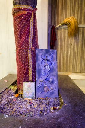 A tree trunk is wrapped in a maroon and gold patterned cloth. In front of the tree trunk, there is a blue artwork depicting a figure with multiple limbs, reminiscent of traditional or religious iconography. Below that, a framed picture rests, featuring colorful imagery. At the base, the ground is scattered with purple-tinted flowers, several small candles, and offerings. To the right, a traditional broom with a long handle leans against a wooden door.