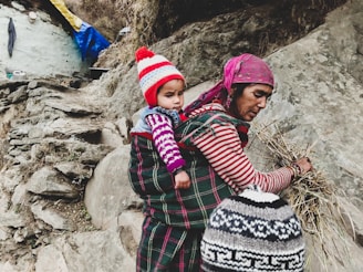 A woman carrying a small child on her back is gathering dried grasses or straw. The woman is wearing a striped long-sleeve shirt and a pink headscarf, while the child is bundled in a colorful, knit cap and sweater. The scene takes place on rocky terrain with what appears to be a rustic dwelling partially visible in the background.
