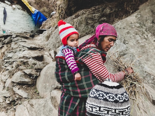 A woman carrying a small child on her back is gathering dried grasses or straw. The woman is wearing a striped long-sleeve shirt and a pink headscarf, while the child is bundled in a colorful, knit cap and sweater. The scene takes place on rocky terrain with what appears to be a rustic dwelling partially visible in the background.