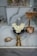 A religious altar features a stone lingam adorned with white flowers, surrounded by brass sculptures including a snake-shaped object, a turtle, and a small vessel. The background displays a decorative statue of an animal.