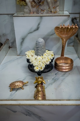 A religious altar features a stone lingam adorned with white flowers, surrounded by brass sculptures including a snake-shaped object, a turtle, and a small vessel. The background displays a decorative statue of an animal.