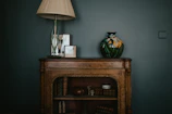 A vintage wooden cabinet is decorated with a tall lamp with an ornate base and a large ceramic vase featuring floral patterns. Several framed pictures are placed next to the lamp. The cabinet shelves hold antique books and decorative items such as a small sculpture and a round container.