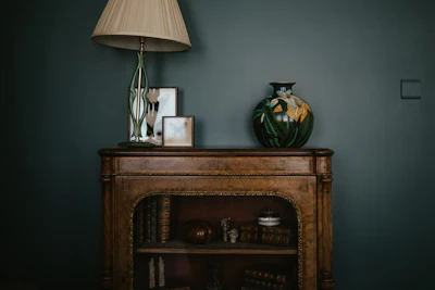 A vintage wooden cabinet is decorated with a tall lamp with an ornate base and a large ceramic vase featuring floral patterns. Several framed pictures are placed next to the lamp. The cabinet shelves hold antique books and decorative items such as a small sculpture and a round container.