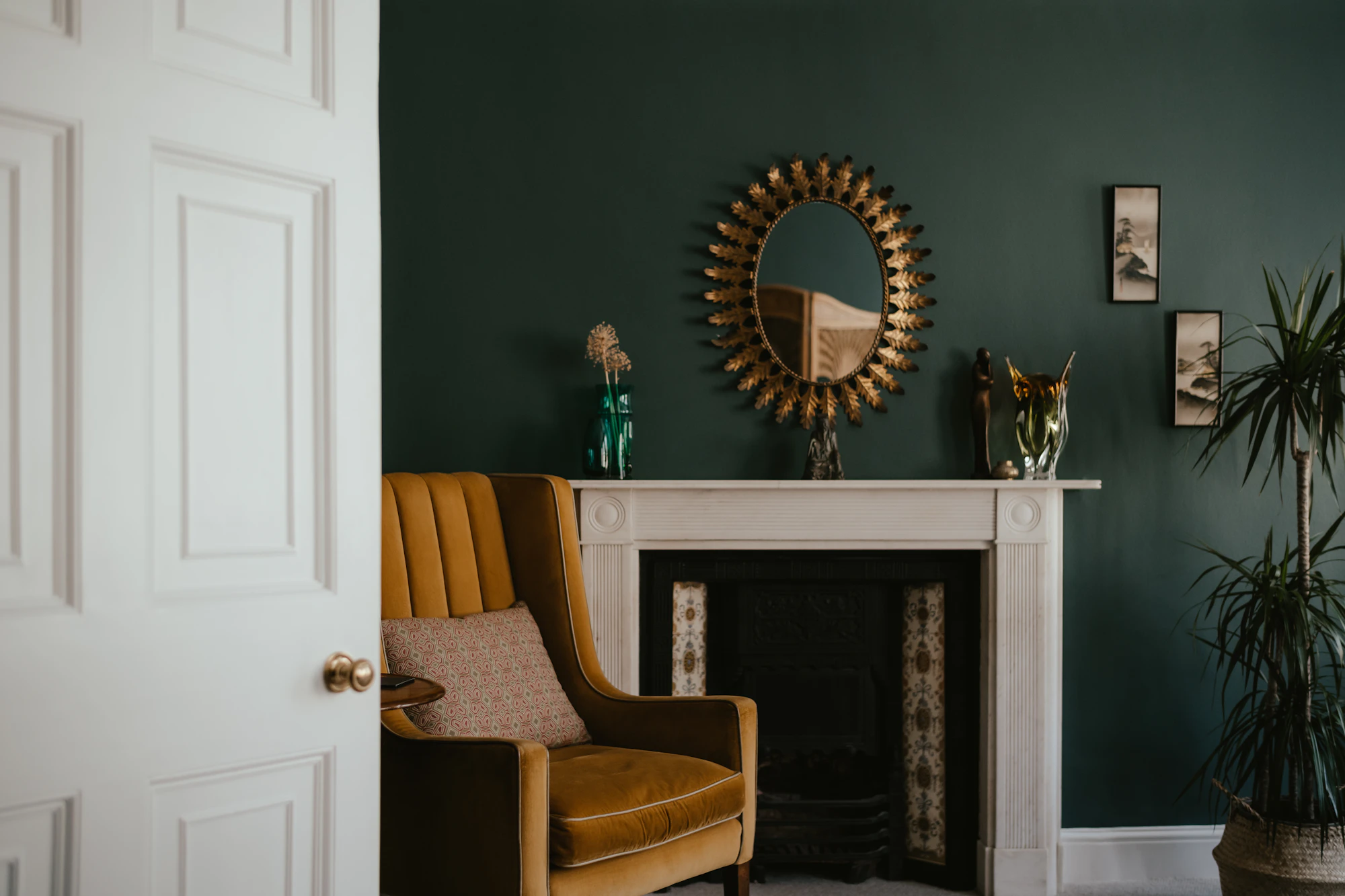 A deep green sitting room: ochre velvet armchair beside a white fireplace, brass mirror above.
