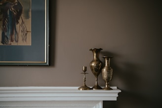 A collection of metallic vases and candle holders is arranged on a white mantelpiece against a muted brown wall. To the left, a framed abstract painting with dark and earthy tones complements the overall subdued color palette.