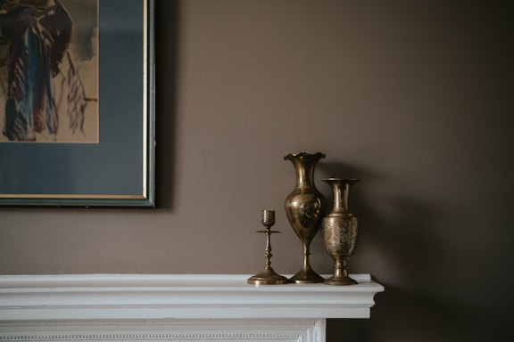 A collection of metallic vases and candle holders is arranged on a white mantelpiece against a muted brown wall. To the left, a framed abstract painting with dark and earthy tones complements the overall subdued color palette.