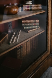 A wooden bookshelf is filled with an assortment of antique leather-bound books, some stacked horizontally and others standing upright. On the shelves are decorative items, including a small sculpture and a vase. The lighting is soft and creates a warm ambiance.