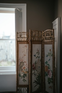 A decorative folding screen stands in front of a softly lit window, featuring intricate floral designs and birds. The screen has a natural brown frame with delicate, colorful artwork including blossoms and foliage. The room has a muted, cozy atmosphere.