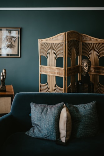 A chic and modern interior scene features a dark teal sofa with two textured pillows in shades of blue and beige. Behind the sofa, a decorative wooden room divider with intricate patterns stands next to a bust sculpture. A wooden table next to the divider holds an abstract metallic sculpture. An artistic framed photograph hangs on the dark green wall, adding to the sophisticated ambiance.