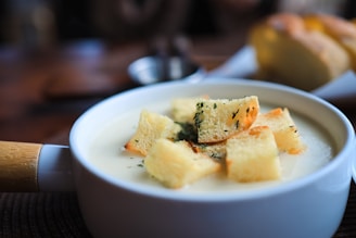 A creamy soup is served in a white bowl with a wooden handle. The soup is topped with several crispy croutons garnished with herbs. In the background, there is a blurred image of a piece of bread on a plate.
