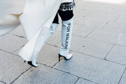A person is walking on a paved sidewalk, wearing high-heeled white boots that have the words 'FOR WALKING' printed on them in black letters. The person is also wearing a light-colored flowing coat and patterned clothing. The scene is bright, with shadows cast on the pale stone tiles of the walkway.