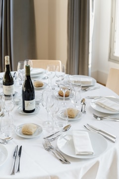 A formally set dining table featuring white tablecloths, white plates with folded napkins, wine glasses, cutlery, and bottles of wine. Small bread rolls are placed on separate small plates. The setting conveys a sense of elegance and preparation for a meal.