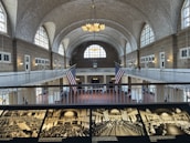 A large, historic hall with a vaulted ceiling featuring intricate brickwork and numerous arched windows allowing natural light. Two large American flags hang prominently in the center. A balcony runs around the upper level, with people visible walking and observing the surroundings. Below, the hall is open and spacious, with historical black-and-white photographs displayed in the foreground.