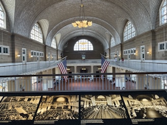 A large, historic hall with a vaulted ceiling featuring intricate brickwork and numerous arched windows allowing natural light. Two large American flags hang prominently in the center. A balcony runs around the upper level, with people visible walking and observing the surroundings. Below, the hall is open and spacious, with historical black-and-white photographs displayed in the foreground.