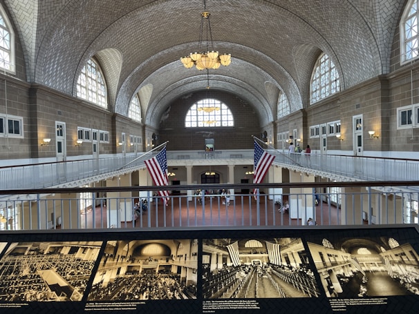 A large, historic hall with a vaulted ceiling featuring intricate brickwork and numerous arched windows allowing natural light. Two large American flags hang prominently in the center. A balcony runs around the upper level, with people visible walking and observing the surroundings. Below, the hall is open and spacious, with historical black-and-white photographs displayed in the foreground.
