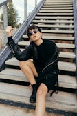A person sitting on outdoor steps, wearing a black leather jacket, sunglasses, and black boots. The style conveys a sense of confidence and modern fashion.