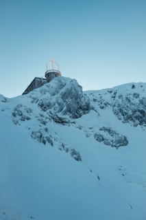 A snowy mountainside with a building at the top, featuring an observatory or antenna structure with a dome and radar equipment. The landscape is rugged with exposed rock formations covered in snow, creating a serene and isolated atmosphere.