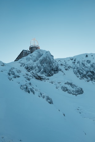 A snowy mountainside with a building at the top, featuring an observatory or antenna structure with a dome and radar equipment. The landscape is rugged with exposed rock formations covered in snow, creating a serene and isolated atmosphere.