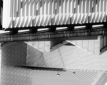 An abstract architectural scene featuring overlapping geometric shapes and patterns. The structures display a mix of diagonal and vertical lines, creating a dynamic visual effect with shadows cast on the surfaces.