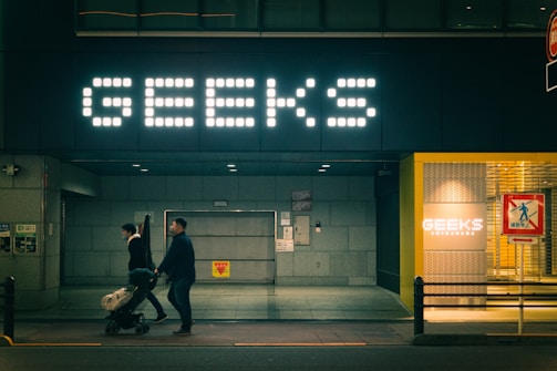 A dimly lit street scene featuring a large illuminated sign with the word 'GEEKS' above a storefront. Two individuals are in motion, pushing a stroller. The entrance is modern, with another smaller sign displaying the same word inside a well-lit area to the right. A colorful pedestrian sign appears in the foreground.