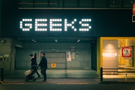 A dimly lit street scene featuring a large illuminated sign with the word 'GEEKS' above a storefront. Two individuals are in motion, pushing a stroller. The entrance is modern, with another smaller sign displaying the same word inside a well-lit area to the right. A colorful pedestrian sign appears in the foreground.