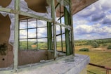 A broken window with shattered glass is in the foreground, with a picturesque countryside landscape visible through it. The landscape includes rolling hills, scattered trees, and fluffy clouds across a blue sky.