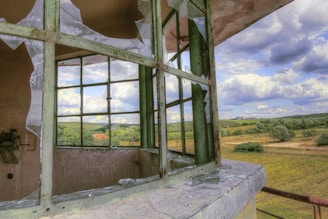 A broken window with shattered glass is in the foreground, with a picturesque countryside landscape visible through it. The landscape includes rolling hills, scattered trees, and fluffy clouds across a blue sky.