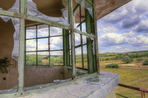 A broken window with shattered glass is in the foreground, with a picturesque countryside landscape visible through it. The landscape includes rolling hills, scattered trees, and fluffy clouds across a blue sky.