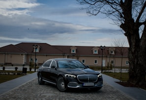A luxury black car is parked on a cobblestone driveway in front of a beige building with a brown roof. The setting includes barren trees and vintage-style street lamps as dusk falls. The sky is overcast, adding to the subdued atmosphere.