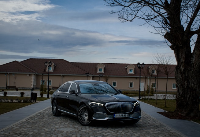 A luxury black car is parked on a cobblestone driveway in front of a beige building with a brown roof. The setting includes barren trees and vintage-style street lamps as dusk falls. The sky is overcast, adding to the subdued atmosphere.