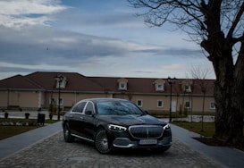 A luxury black car is parked on a cobblestone driveway in front of a beige building with a brown roof. The setting includes barren trees and vintage-style street lamps as dusk falls. The sky is overcast, adding to the subdued atmosphere.