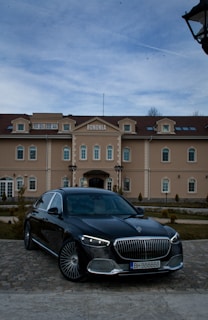 A sleek black luxury car is parked on a cobblestone driveway in front of a large, elegant beige building with a red-tiled roof. The building is symmetrically designed with multiple windows and decorative elements. It has the word 'BONONIA' on the facade. The sky above is lightly overcast, suggesting a calm day.